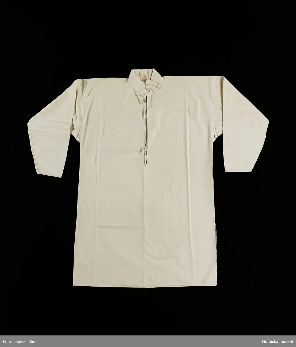 Mansskjorta  i traditionell modell, av oblekt lakanslärft i bomull, ett bålstycke med sidsömmar och sprundi sidorna. Rak ståndkrage med öppet sprund , knyts med bomullssnodd i halsen. Vidsydd rak ärm med ärmspjäll, inga ärmlinningar. Helt sydd för hand.
Skjortan hör uppenbart till de plagg som sytts upp speciellt för Nordiska museet på beställning av Artur Hazelius. Beställningarna ombesörjdes av handlaren Söderman som lät kunnigt folk i bygden sy efter gammal modell.
/Berit Eldvik 2010-09-14