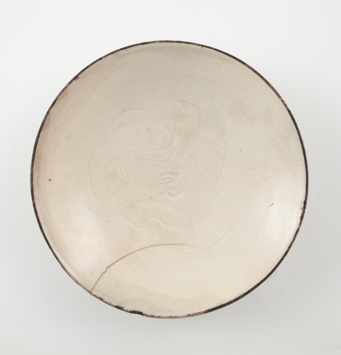 Lav, rund porselensskål med kremhvit glasur. Innrisset et slyngplantemønster i speilet. Rester etter kobberkant.