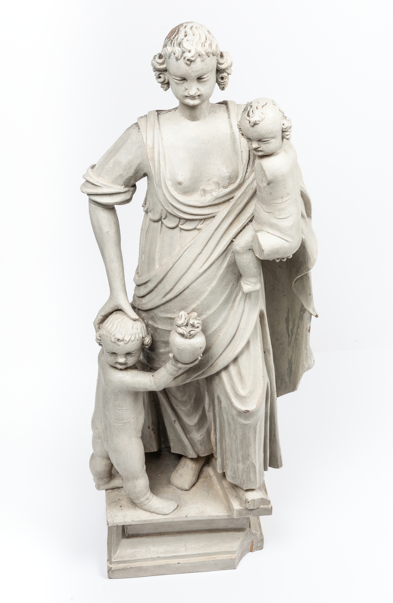 Kyrklig skulptur, trä, staty från gamla korskranket i Heliga Trefaldighets kyrka i Gävle, av Ewert Friis omkring 1665. 

Stående kvinnofigur med naket barn på vänster arm samt stående naket barn vid höger sida. Sistnämnda barn har ett brinnande hjärta i höger hand och kvinnans vänstra hand på sitt huvud. Vänster ben är främst, foten avslagen. Sittande barnets ena ben och andra fot avslagna. 
Fyrsidigt, profilerat postament. 
Gråvit färg.
