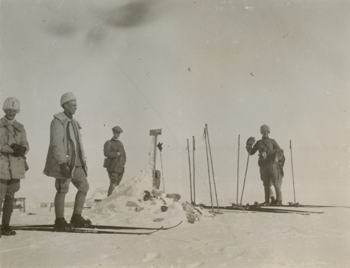 Grupp soldater och en civilkläd man vid ett stenröse på fjället. Sidor ligger på marken och stavar är stuckna i snön.