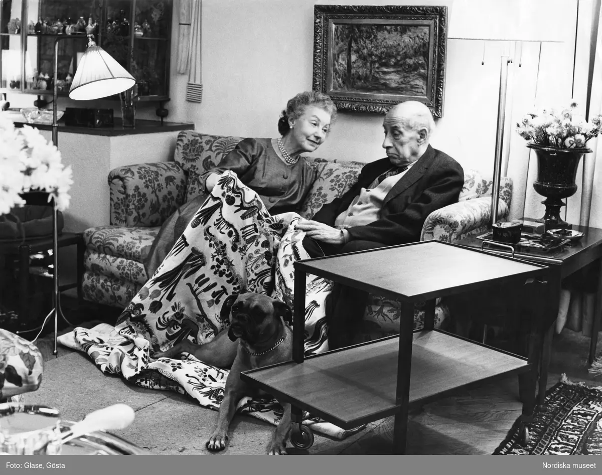 Svenskt Tenns grundare Estrid Ericson och hennes vän och medarbetare arkitekten Josef Frank sitter tillsammans i en soffa och tittar på tyget Primavera som han formgivit. Med på bilden är Estrids boxerhund.