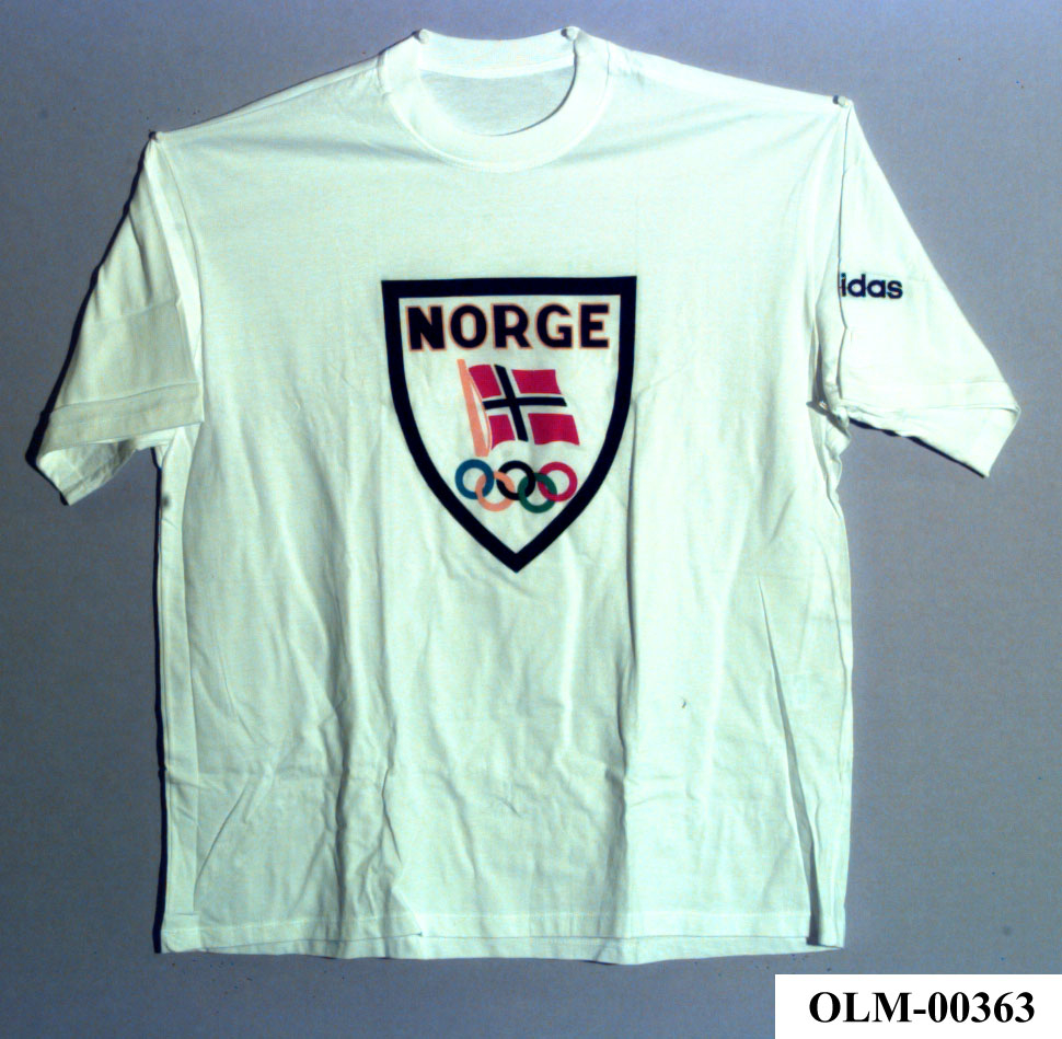 Hvit T-skjorte fra Adidas med Norges Olympiske Komités emblem på brystet. Emblemet er formet som et skjold med svart kant, teksten Norge, det norske flagget og de olympiske ringer.