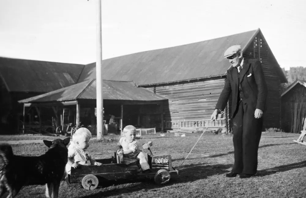 Svart-hvitt foto avg mann i dress og sixpence som drar en olabil med to barn på, Tomterlåven i bakgrunnen.