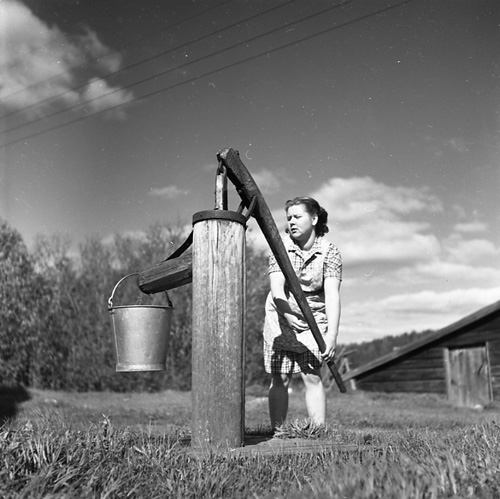 En kvinna står på gårdsplanen vid Sörgården i Västerböle och pumpar vatten från vattenpumpen in i en hink.