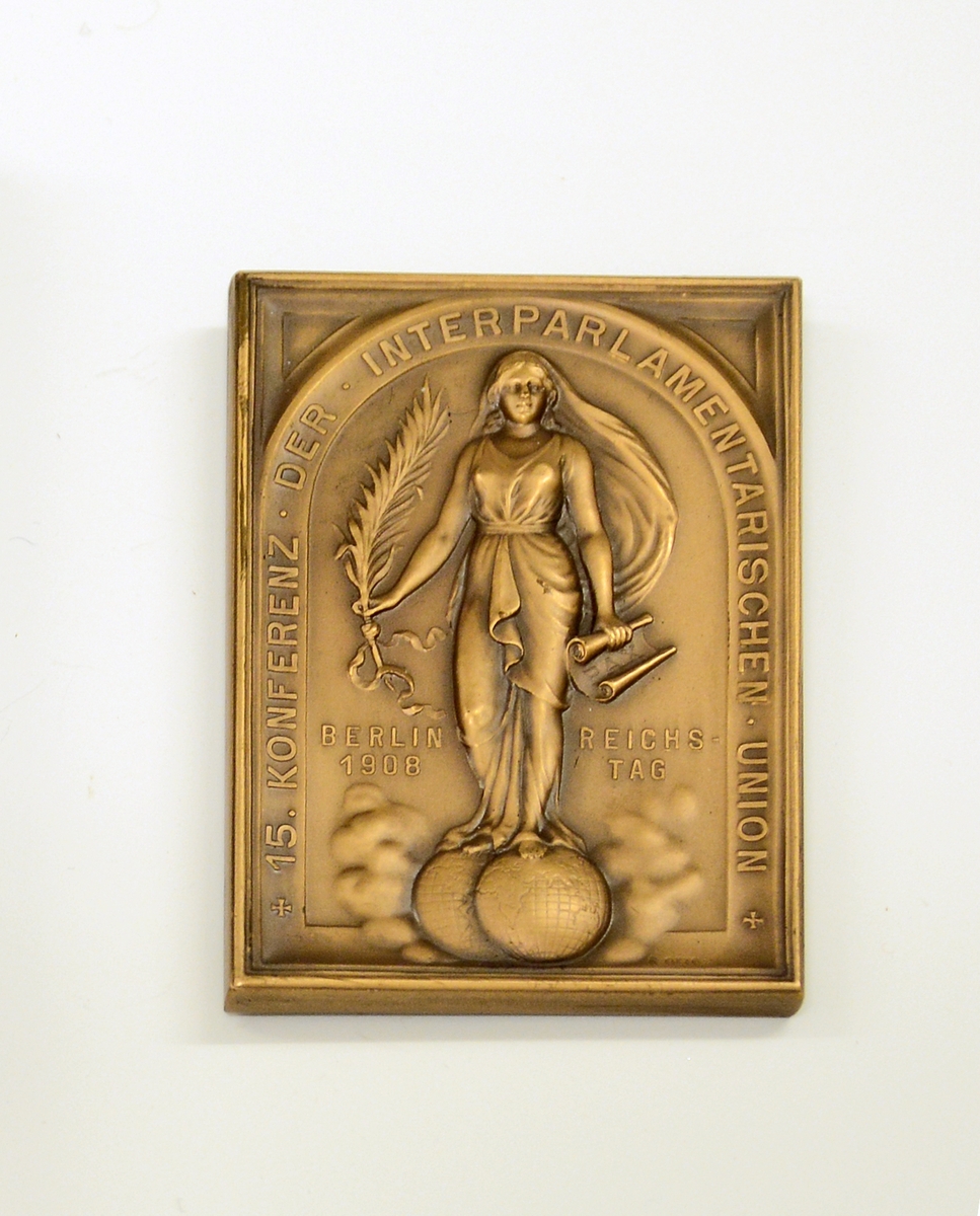 Fra protokollen: 
Plate i bronse, i etui. Platen er rektangulær, med målene 6,8 cm x 4,7 cm. På forsiden istøpt bilde av keiser Wilhelm II, hans navn og "DEUTSCHER KAISER KØNIG V. PREUSSEN" under bildet. På den andre siden bilde av en gudinne (?) som står med en globus under hvert ben, i venstre hånd en dokumentrull med ordet "PAX" på, i høyre holder hun en plantegren. Rundt figuren står følgende:   "+ 15. KONFERENTZ - DER - INTERPARLAMENTARISCHEN - UNION +". 
Ved siden av figuren: "Berlin 1908" "Reichstag".
Etuiet har sort imitert skinntrekk og på forsiden står: "Interparlamentarische Union XV. Konferentz. Berlin 1908. Reichstag". På innsiden av lokket foret med grønn silke. I gullbokstaver står: "R. OTTO. Kgl. Hofgraveur. Berlin. U. d. Linden 40." Bunnen er innvendig trukket med mørk grønn fløyel.