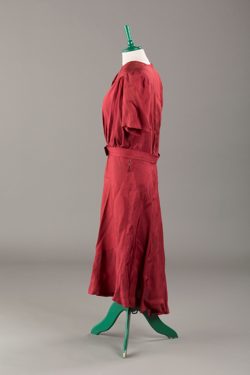 Form: Glatt, klokkeformet skjørt. Forstykkene rynket til buede bærestykker foran. Overdel rynket til skjørt. Korte ermer. Lukket med trykknapper. Belte fastsydd.