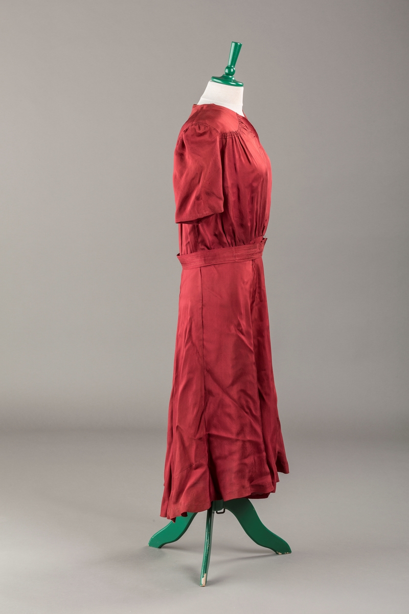 Form: Glatt, klokkeformet skjørt. Forstykkene rynket til buede bærestykker foran. Overdel rynket til skjørt. Korte ermer. Lukket med trykknapper. Belte fastsydd.