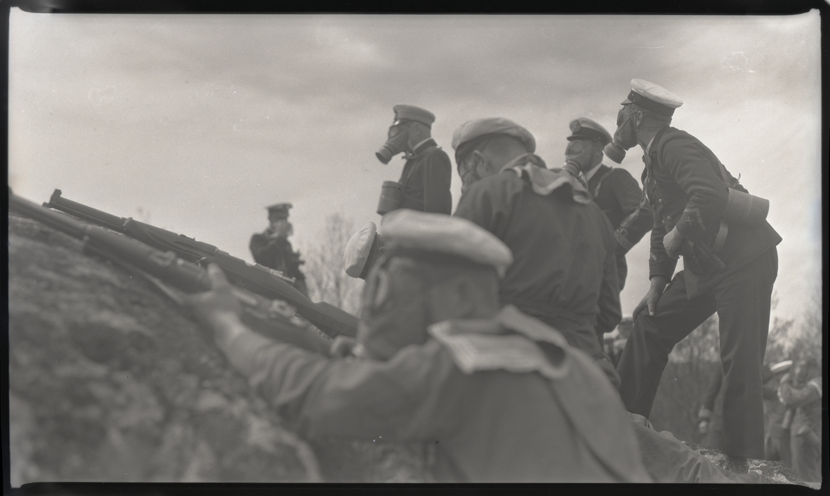 Officerare och manskap iförda gasmask under stridsövning. Av andra bilder i Bergers samling framgår att denna bild tagits under landstigningsövning med besättningen från pansarskeppet SVERIGE på Stabbo i Stockholms södra skärgård 1929.