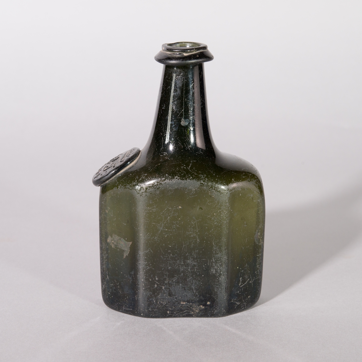 Flaska av grönt glas. 8-kantig kropp, axlar och rund hals. Sigill.