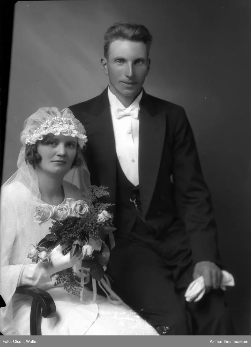 Ateljébild på ett brudpar. Kvinnan har brudklänning och slöja samt blombukett. Enligt Walter Olsons journal är bilden beställd av Enoc Abrahamsson.