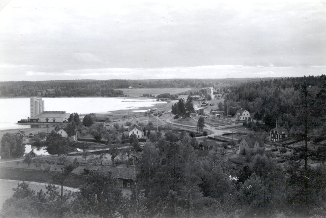 Vid Östersjöns strand i viken Syrsan ligger Helgenäs. Bilden visar en utsikt över orten. Bland annat syns en anläggning med silobyggnad, uppenbarligen anlagd med kajplats.