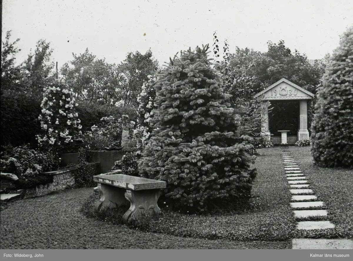 1930-monumentet i Widebergs trädgård.

John Wideberg blev tidigt intresserad av
trädgårdsanläggningar. Han hade sannolikt hämtat
inspiration på resor han gjort till Stockholmsutställningen
1897 och till Göteborg 1923. Han blev också vän med
stadsträdgårdsmästaren Haglund i Kalmar.
John hade palmer i potatiskällaren under huset och påfåglar,
ekorrar och pärlhöns som övervintrade i hönshuset. En
papegoja och en sköldpadda fick tillbringa vintrarna inne i
köket. Runt trädgården planterade han skyddande häckar.
Arbetet påbörjades redan när han var 18 år med att han
hämtade åtta granar i skogen, och planterade dem i en
ring mitt i trädgården. Dessa finns kvar än idag. I slutet
av 1930-talet arrenderades jordbruket ut och John kunde
ägna sig på heltid åt trädgården, som blev alltmer av ett
besöksmål. John började ta inträde, 25 öre.
John utvecklade också sin hobby som tavelmålare och som
fotograf. Han tog med sin lådkamera under årens lopp
hundratals vackra bilder från trädgården, liksom på vyer,
hus och människor i takten. Ca 500 av dessa bilder finns
bevarade som glasplåtar, skänkta till Kalmar läns museum.