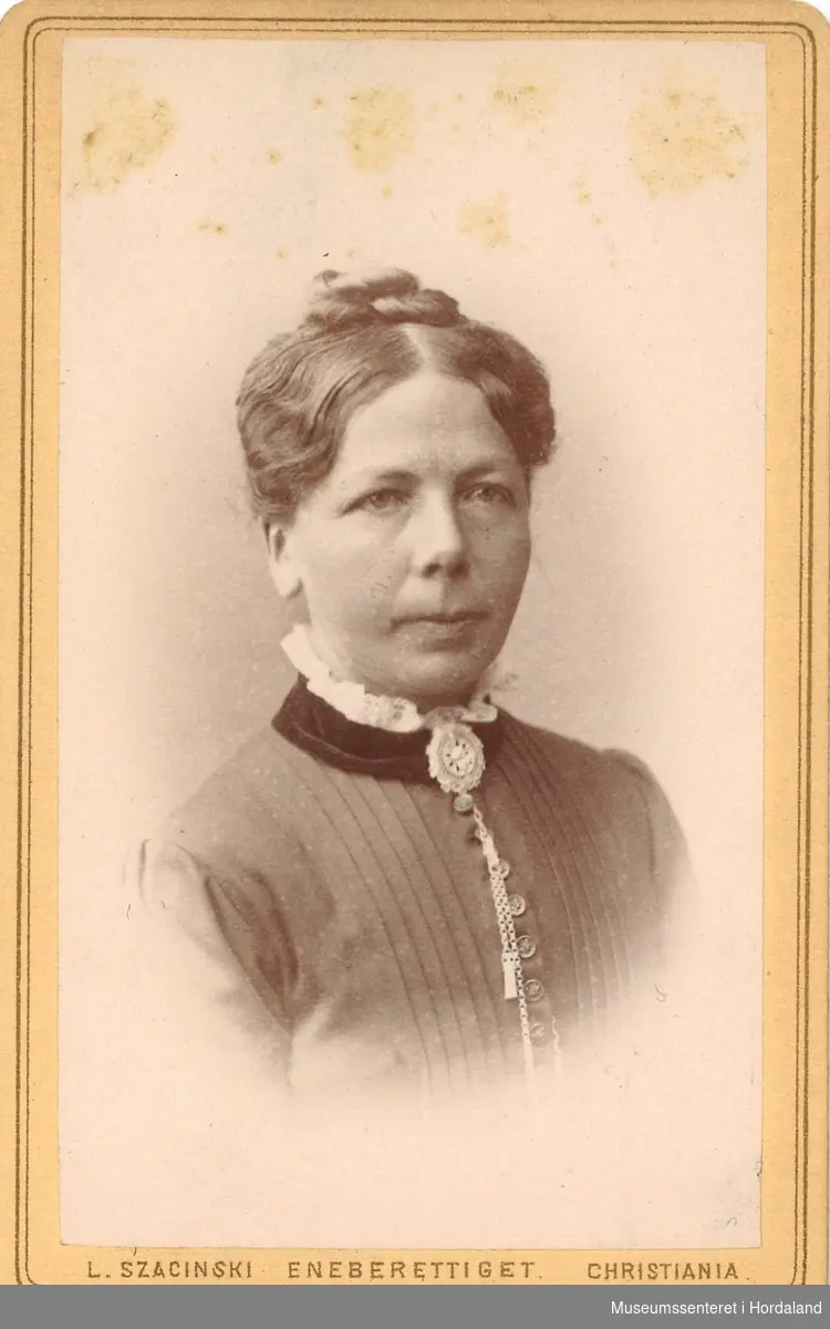 halvfigur fotografi av kvinne med hår med midtskill og topp, kledd i plissert bluse/jakke med brosje i halsen og kvit rysjekrage