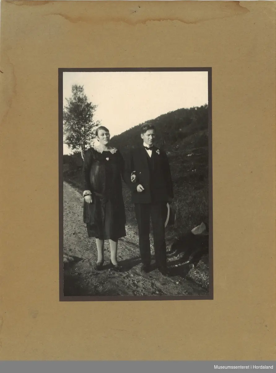 amatørfotografi av kvinne og  mann i mørke festkle som står på ein grusveg med grasbakke og ei enkel bjørk i bakkanten