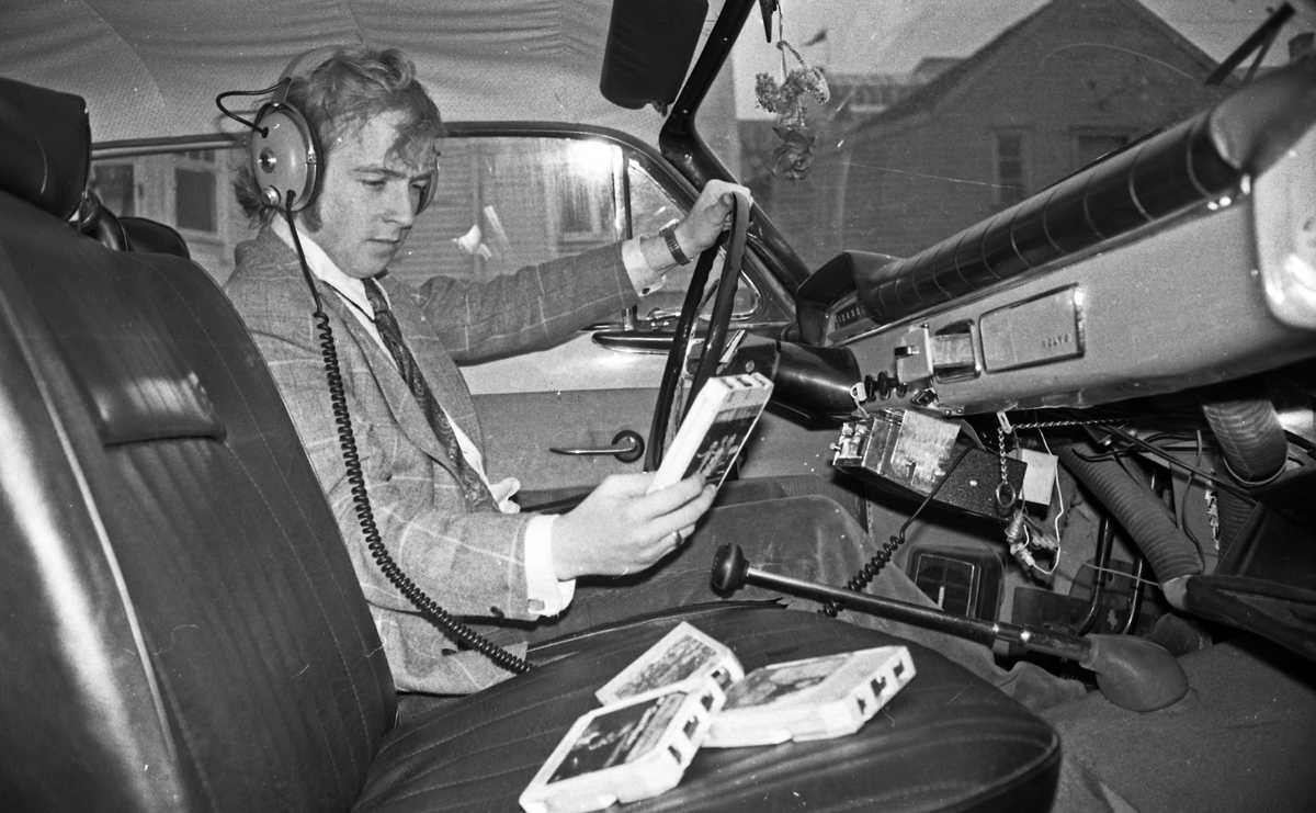 Mann med høreklokker. Testing av stereo i bil. Dyp konsentrasjon. Flere musikk-kassetter liggende på passasjersetet. Bilseteoanlegg av typen 8-spors. (Utdatert i dag i 2015.)