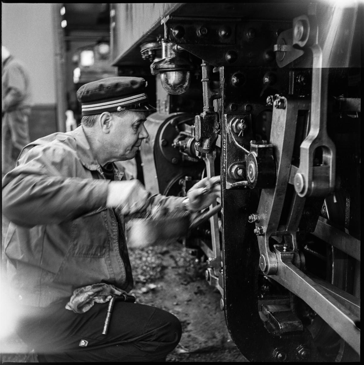 Sort-hvitt fotografi av mannlig frivillig i tjeneste. Står på kne foran understellet på et damplokomotiv. Han har uniformslue på hodet, og oljekanne i den ene hånden.