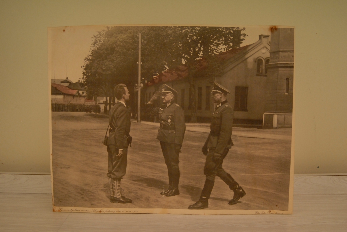 To tyske offiserar i uniform og ein mann i nikkers og rosete knesokkar som helsar på kvarandre.
