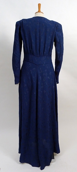 Enligt liggaren: Klänning dam av cloque, blå v-ringad, aftonklänning, draperad vid axlarna, blixtlås i sidan. Lång ärm axelvaddar. Midjebesparing, l 160 cm, bredd över axel 41 cm. 1940-tal