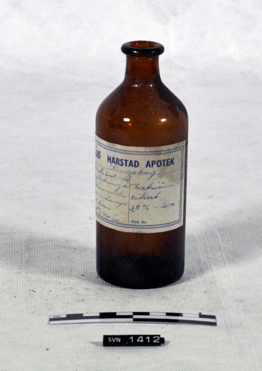 Brun flaske med hvit etikett med håndskrevet innholdsbeskrivelse. Etiketten har trykket logo og navn.
Flasken er tom og har ikke kork