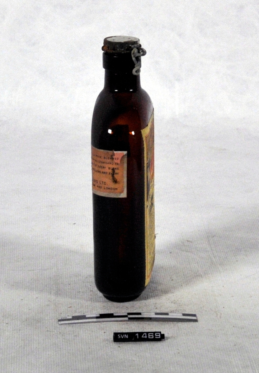 Brun flaske med kork og etikett.
Korken er festet til flasken med hengsel i metall.
Flaskens form er en tynn oval