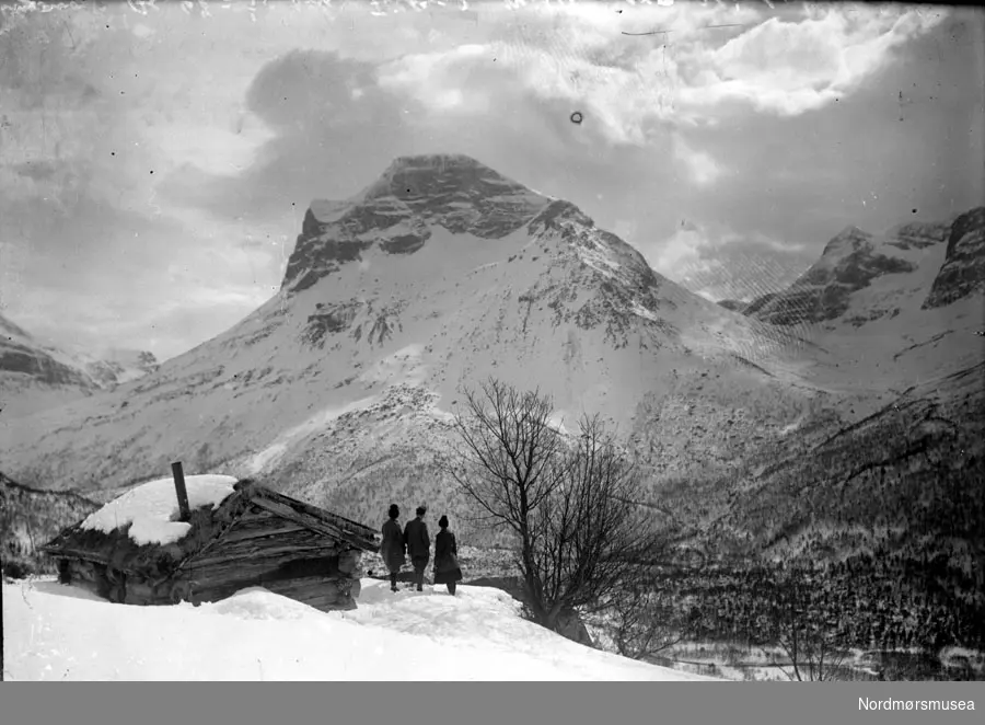 Vinterlandskap på fjellet, med tre mennesker stående utenfor en fjellhytte. Datering usikker. Fra Nordmøre museums fotosamlinger, Myren-arkivet.
