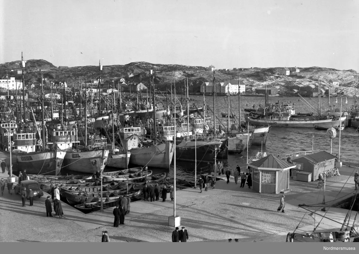 Foto fra Sildeflåten til kai ved Piren/Vågekaia ved havna på Kirkelandet i Kristiansund. Bildet er datert 1954. I bakgrunnen ses Gomalandet. Fra Nils Williams fotoarkiv. Fra Nordmøre museums fotosamlinger.