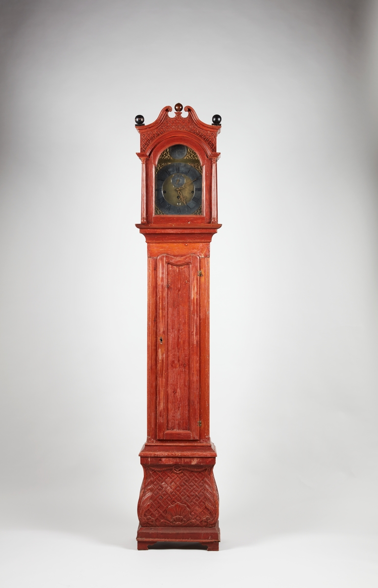 Rødmalt gulvur med forsiringer i form av nettverk, sped, stilisert akantus og rocaille. Urverket har sekund-, minutt- og datoviser og er forsynt med spillverk med to melodier.