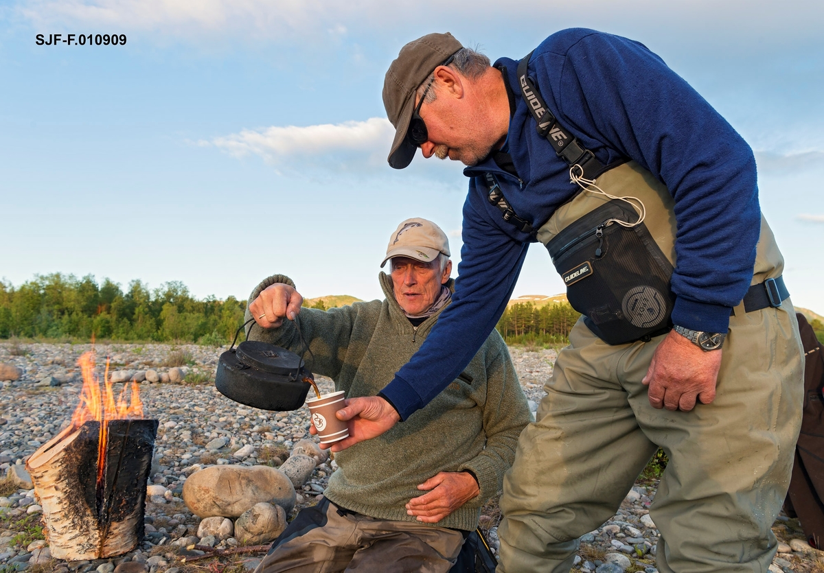 Idar Edvardsen fra Bjerkvik (opprinnelig fra Grovfjord) serverer kaffe fra svartkjele til Walter Hjellvik fra Trondheim. I bakgrunnen en finneprimus eller stubbfyring som han har brukt til kaffekoking. Friluftsliv. Leirliv. Kokekaffe. Kaffe. Bildet er tatt ved Altaelva i Øvre Alta, Finnmark. Idar har nettopp landet en laks på 15 kilo i Altaelva, og det var god stemning i leiren. Laksefiske i Altaelva, Alta, Finnmark. Fiske etter laks. Sportsfiske.