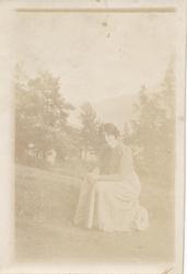 Kvinne sitter ute i hverdagsklær.  Langt skjørt og rutete bl