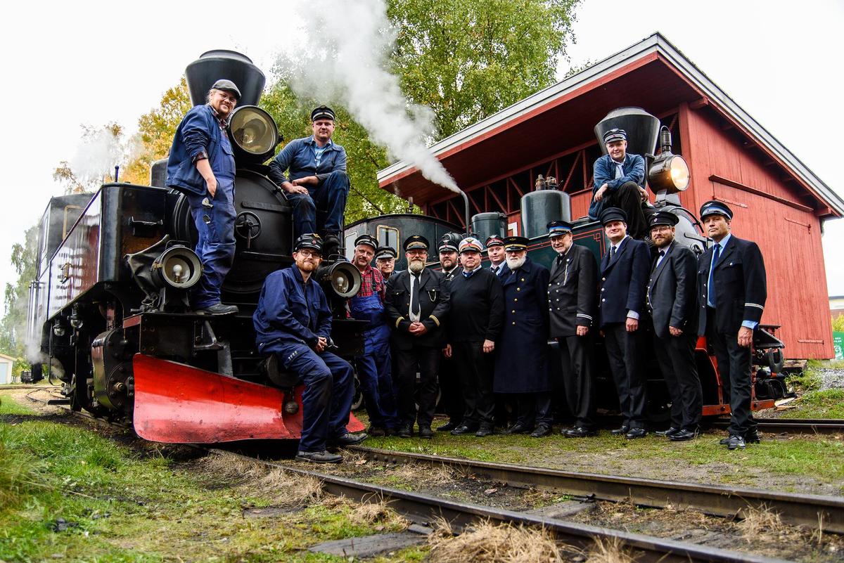 En gruppe mannlige frivillige fra Tertittens venneforening poserer foran et av Urskog-Hølandsbanens damplokomotiv.