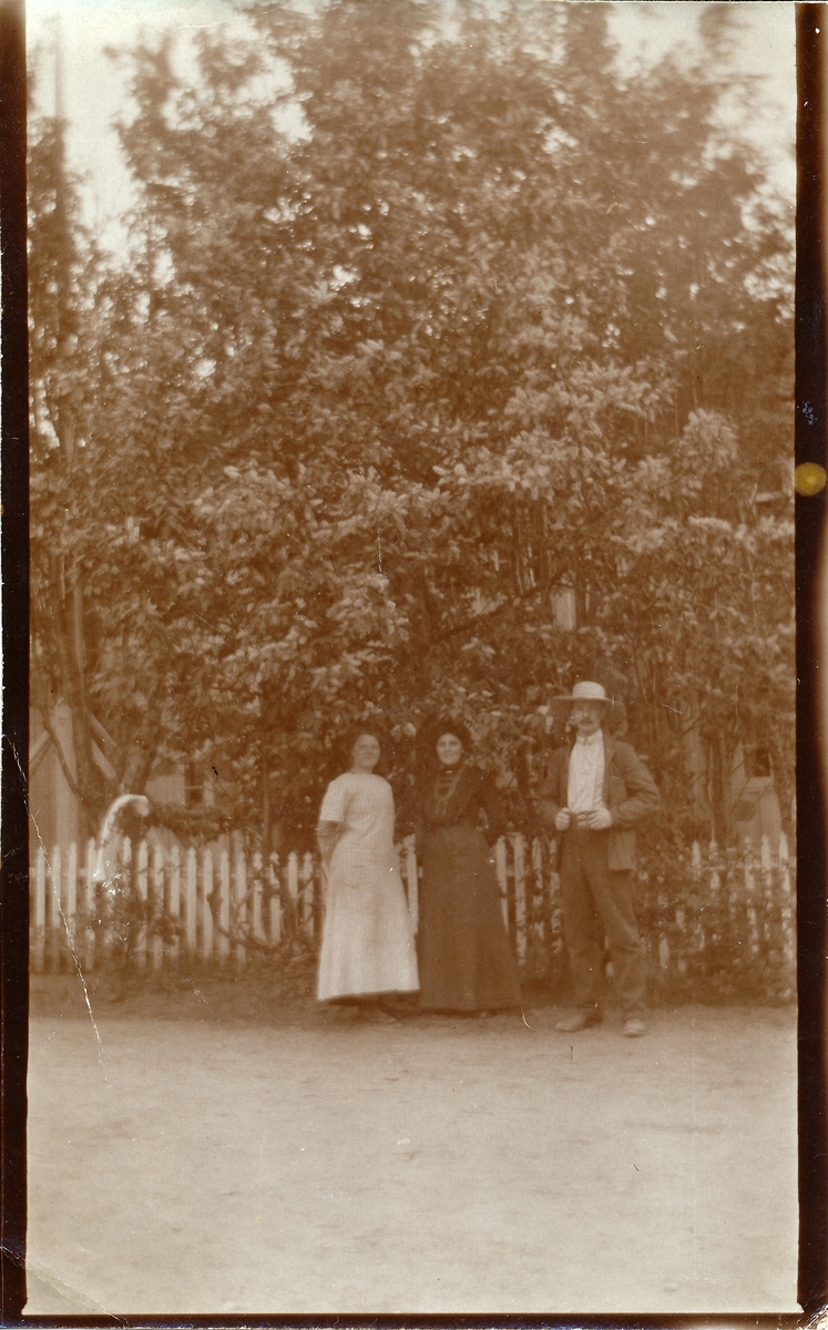 To kvinner og en mann foran stakittgjerde med bygning og trær bak.