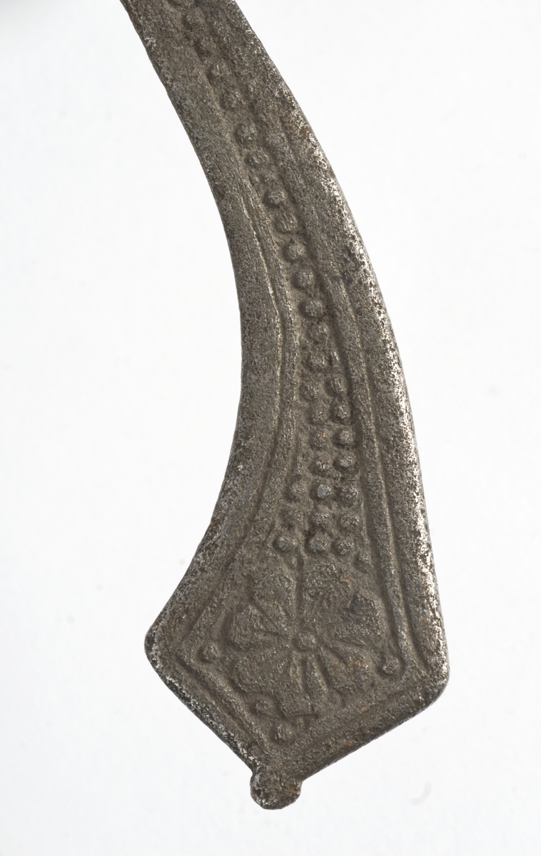 Rund pæreformet knapp med 10 fasetter  Tregrep. Flate svakt opp og nedbøyde  parerstenger med som ender i to fliker. Punslet dekor på parerstengene.
Asymetrisk kurvfeste med nyreformet parerplate på innsiden Prereplaten på utsiden er borte. Sidebøyler og håmdbøyle er formet av flate bånd. Skade på bånd på utsiden.
Tveegget rett klinge med  ovaklt  tverrsnitt  og to korte blodrender. Klingen har innskripsjon "SAHGON"