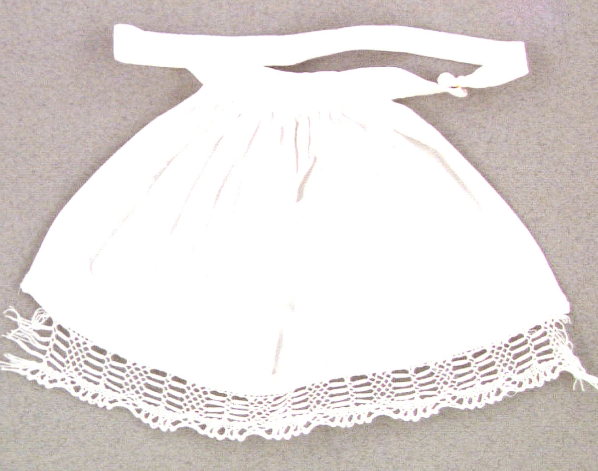 Dockförkläde av vitt bomullstyg med påsydd knypplad spets i nederkanten. Ett linningsband runt midjan som fästes med en tryckknapp