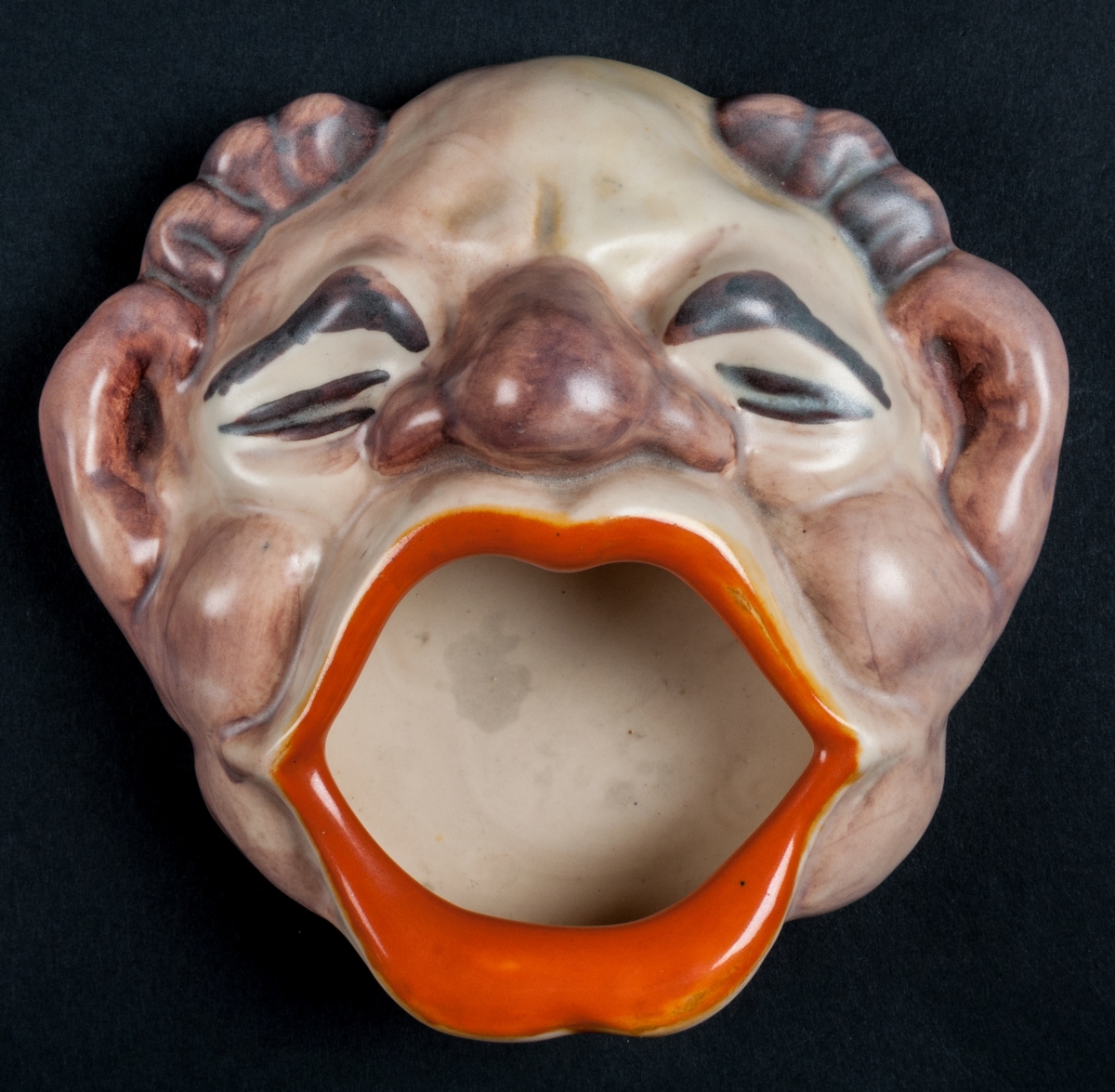 Askfat i form av ett clownhuvud, i keramik. Tillverkad vid Bobergs Fajansfabrik i Gävle 1944-1953. Formgivare: Eva Jancke-Björk.
Märkt: 3506 Bo FAJANS GEFLE.