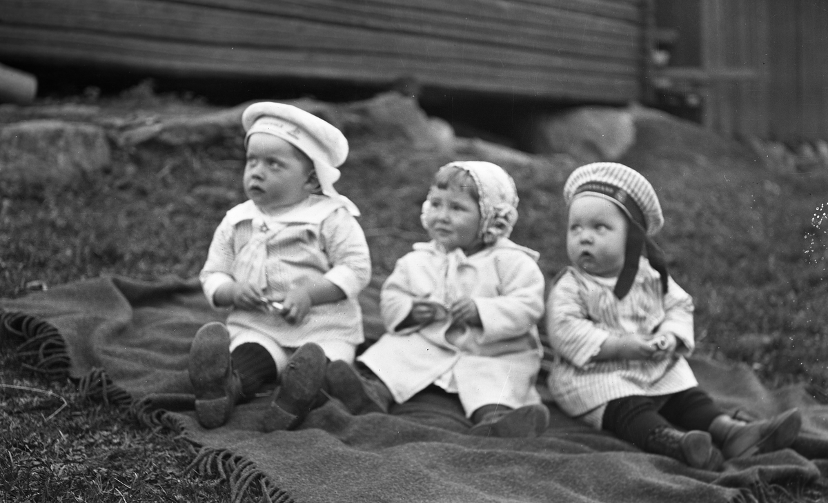 Tre små barn sitter på et teppe på bakken. Fra venstre Anders Holmstad, Nora Gunnerød, og Ole (Olsen) Gunnerød.