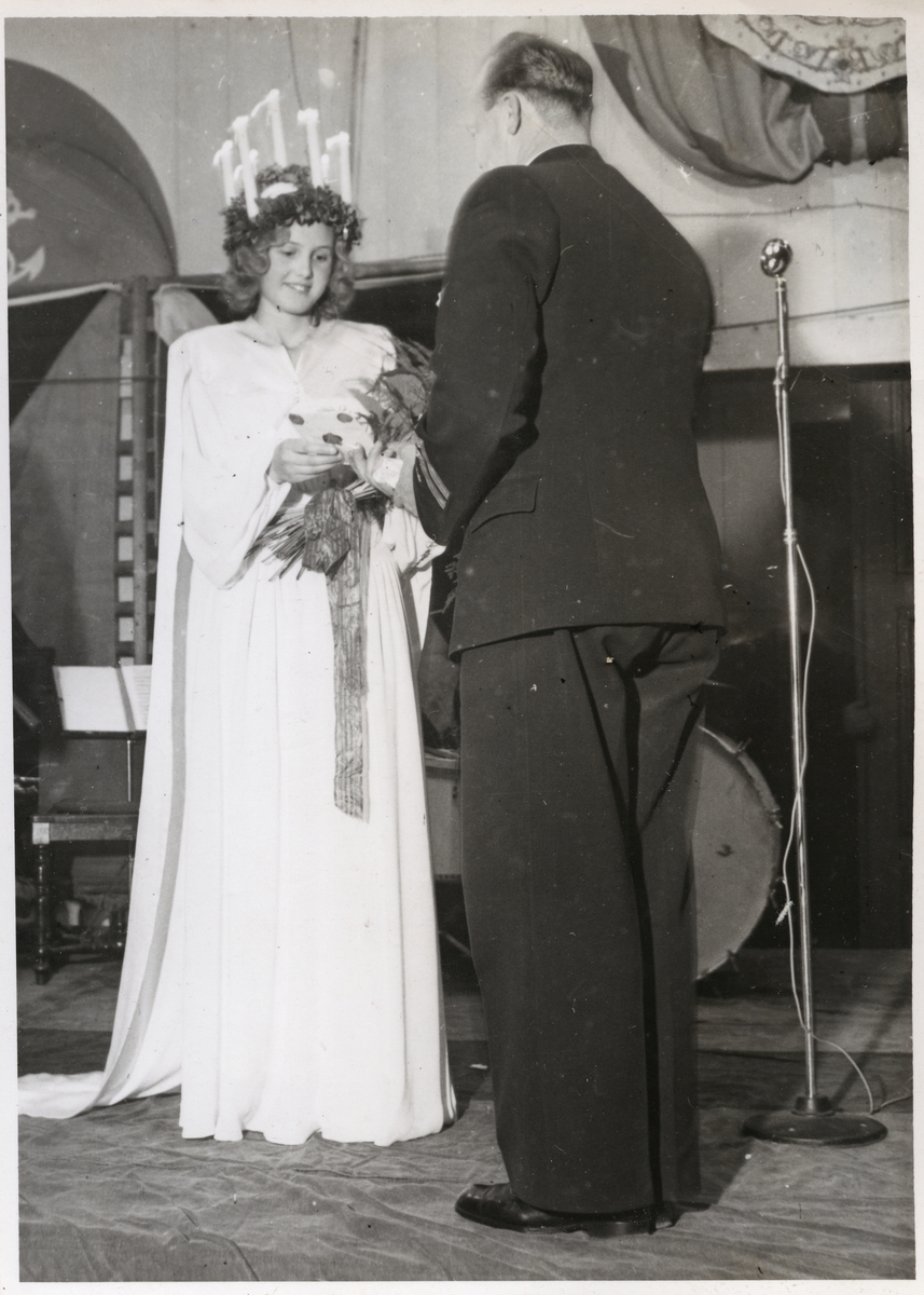 Årets lucia klädd traditionsenligt med ljus i håret mottager en bukett blommor av man med ryggen vänd åt kameran under luciafirande 1943. I bakgrunden syns instrument och mikrofonstativ.