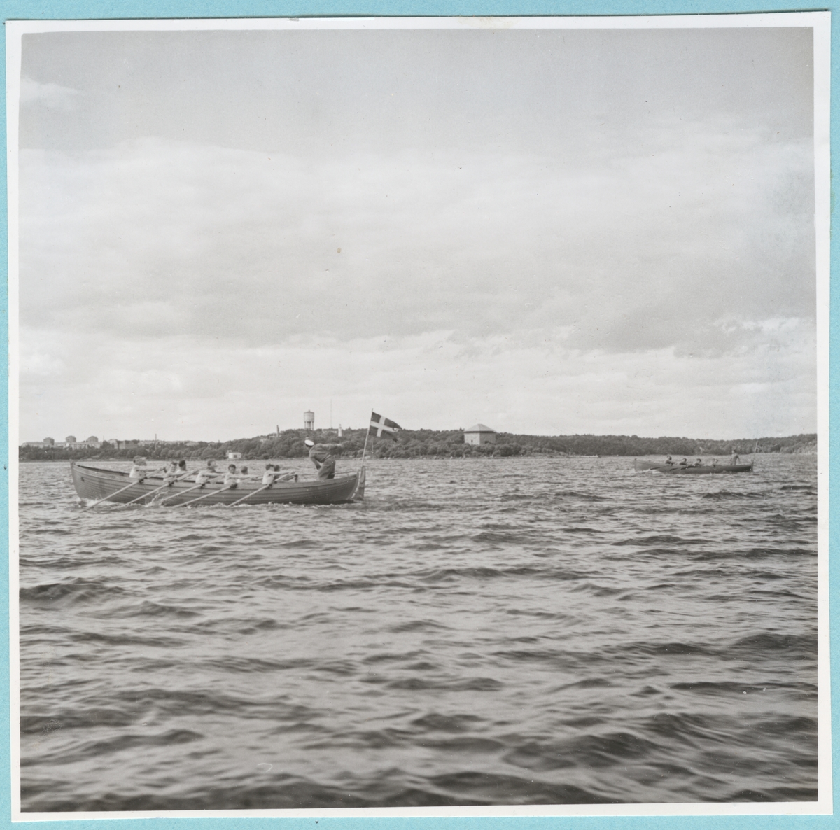 Roddtävling mellan Danska, Norska och Svenska kadettlagen. Två båtar syns ro fram över vattnet. I bakgrunden syns Mjölnareholmen med kruthuset, samt vattentornet på Vämö längre bort. Från Danska och Norska örlogsbesöken den 29/6 - 4/7 1951.