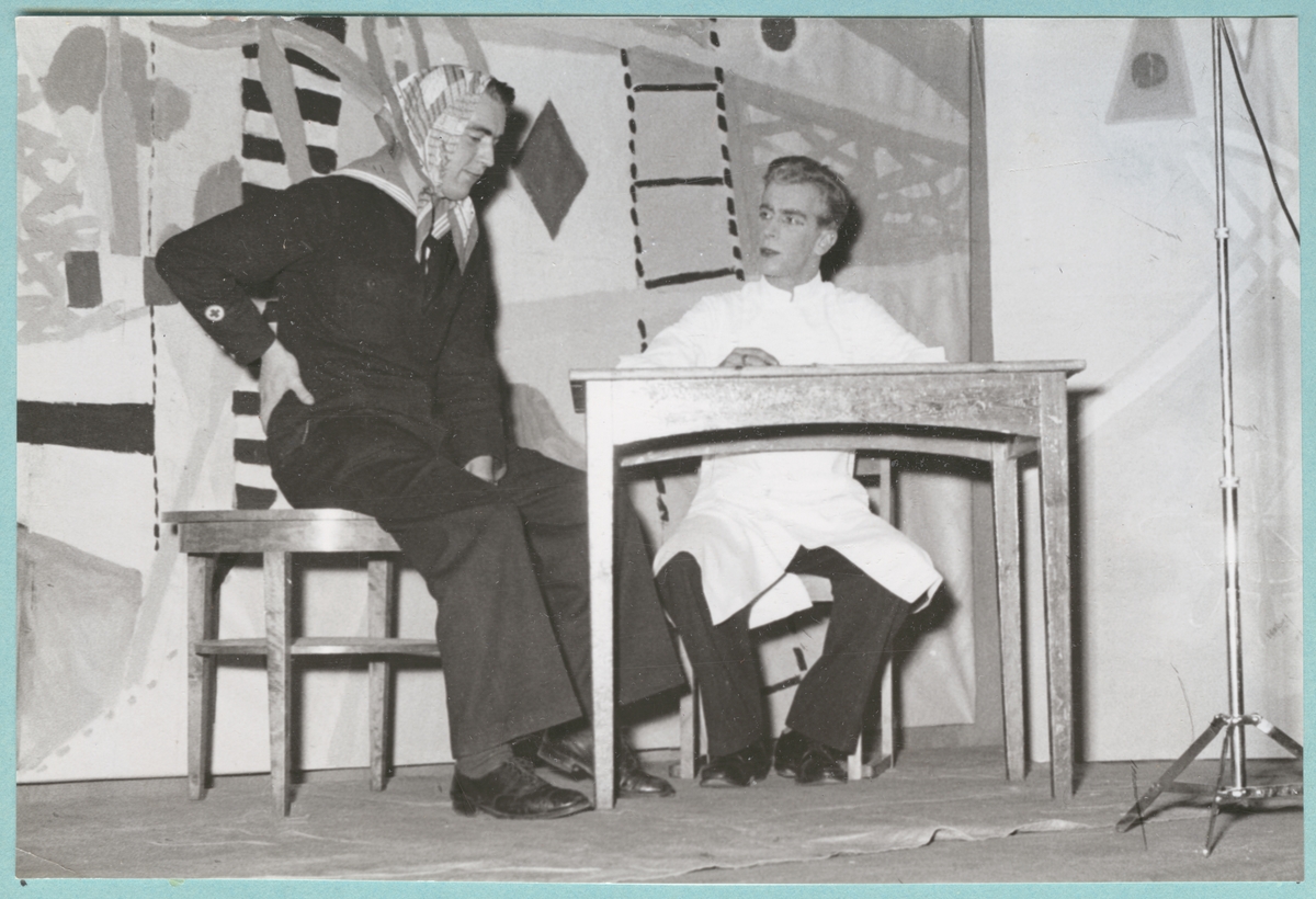 Två utklädda flottister uppträder i en revy på en scen, sittandes framför ett bord. Bakom dem syns en målad bakgrund med färger och former inspirerade av marinen.
