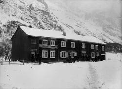 Personer foran ei lån, to hester med slede. Stuebygninga i N