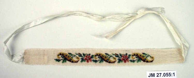 Ett strumpeband, tillhörande ett par, broderat, brodyren på glesvävt vitt bomullstyg är fäst på sidenband. Blomstergirlang av pärlor med blommor i olika färger, troligen använt och broderat av bruden själv.