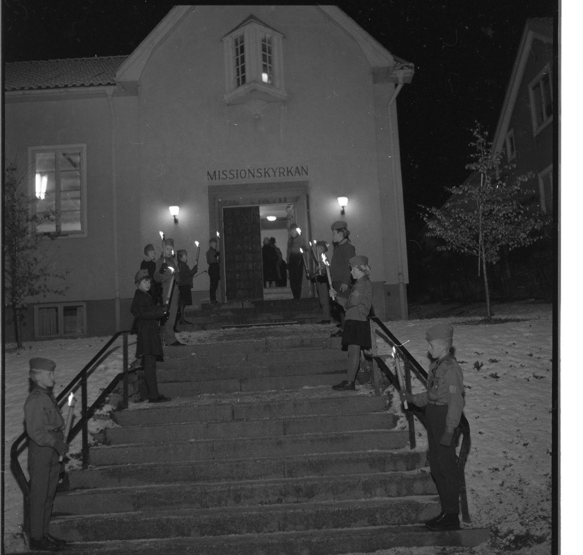 Ungdomar (Scouter/GU?) med facklor placerade vid trappan upp till Missionskyrkan i Gränna.