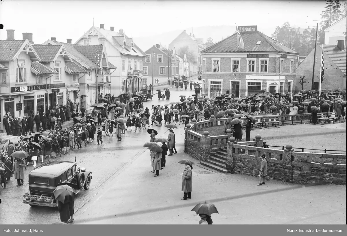 17. mai feiring 1931. Folketog i gaten ved Notodden torg. Tilskuere på fortau. Biler og mennesker gående i gaten. Teledølens lokale i bakgrunnen. Butikkfasader langs Storgata i Notodden sentrum.