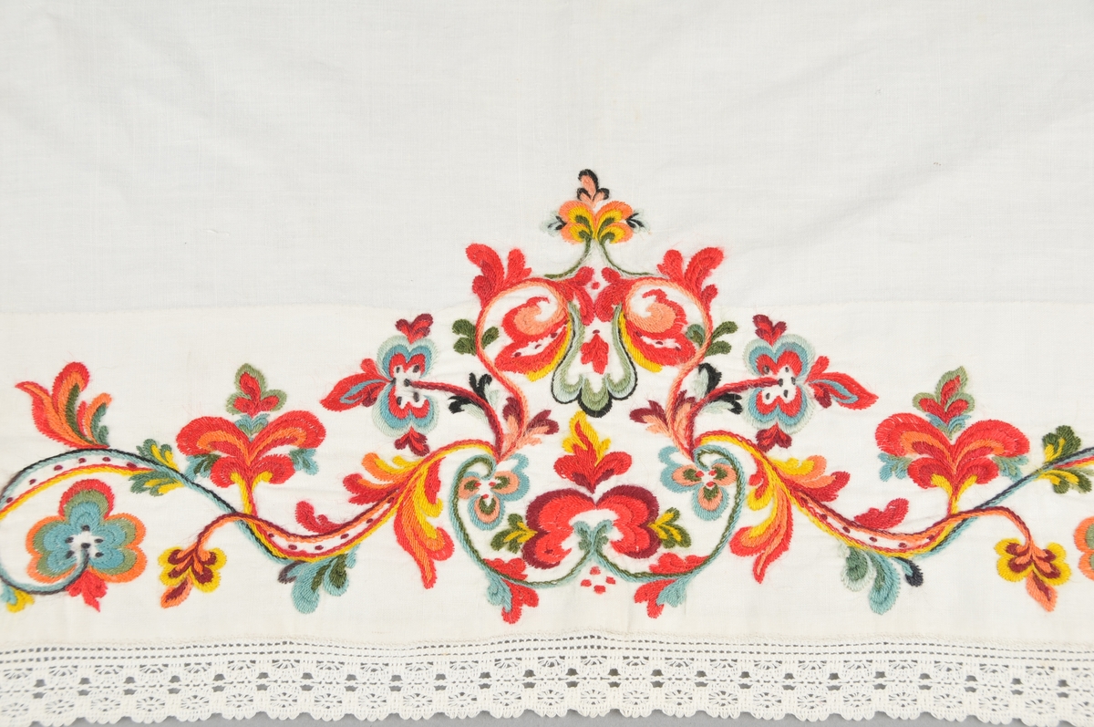 Kinnlag i kvitt linlerret, utsauma med ullgarn ("kamelgarn"), i raudt, grønt, gult, blått, orange og svart. Borden er sauma gjennom dobbelt tøy. Påsydd kvit bomullsblonde på tre sider, jarekant på kortsidene og falda på langsida.