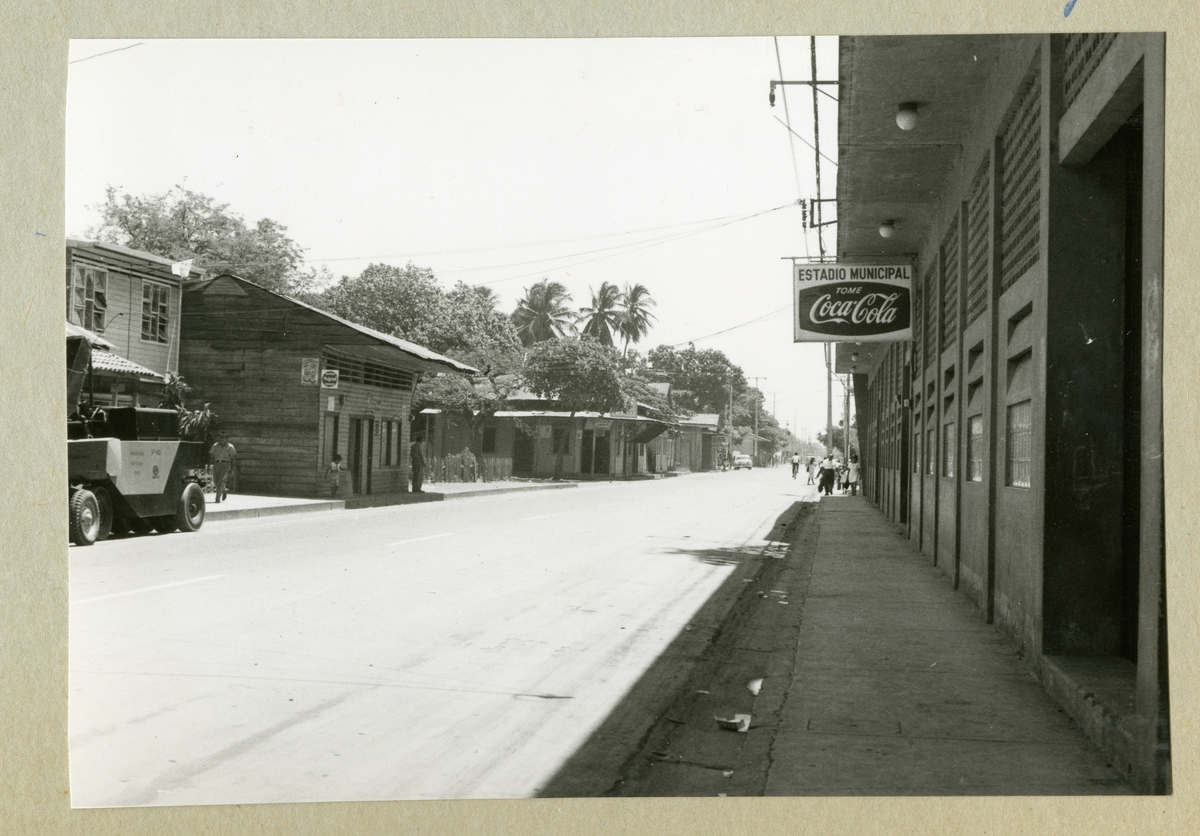 Bilden föreställer en gata i Puntarenas som kantras av växtlighet och mindre byggnader. På en av byggnaderna syns en reklamskylt för Coca-Cola mycket tydligt. Bilden är tagen i samband med minfartyget Älvsnabbens långresa 1966-1967.