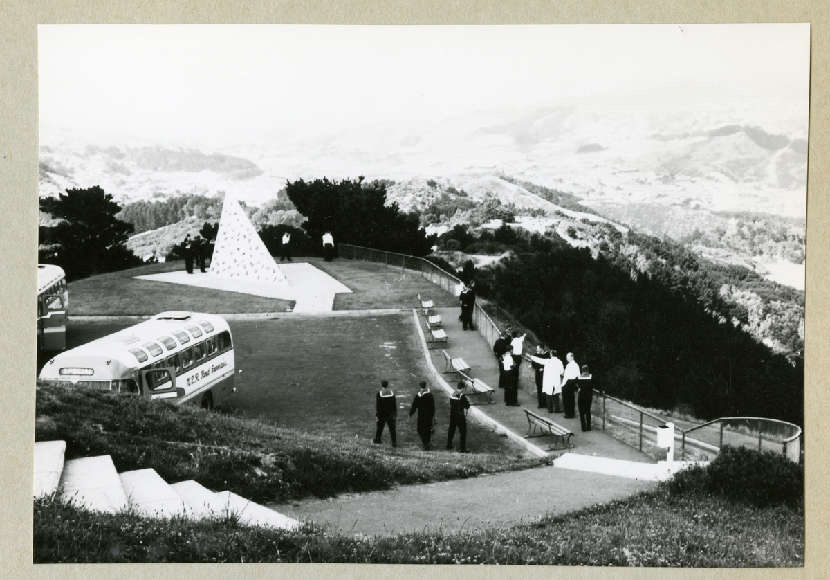 Bilden föreställer ett landskap med kullar och dalar. Vid en gräsplätt syns en rad bänkar, två bussar och uniformsklädda män. Bilden är tagen under minfartyget Älvsnabbens långresa 1966-1967.