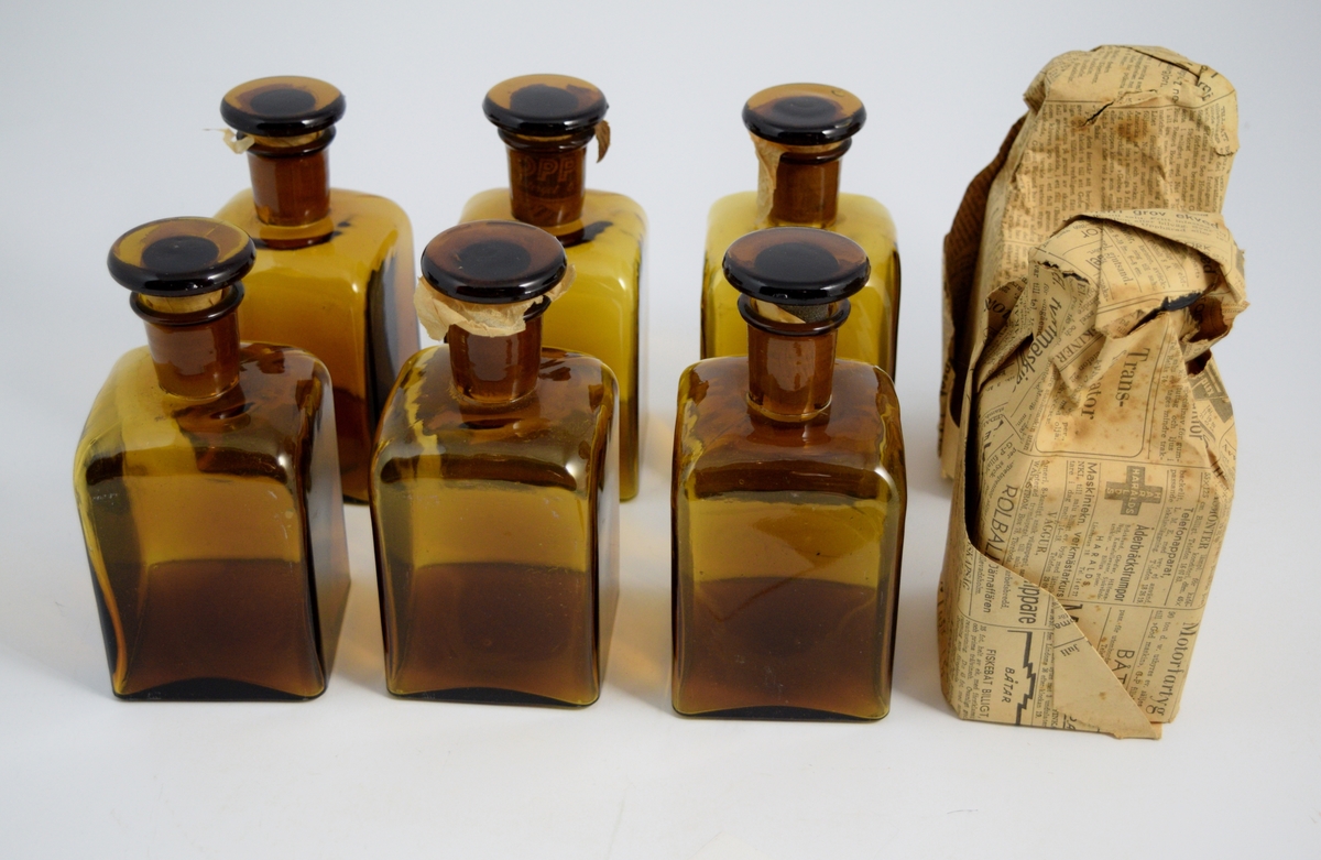 Brun firkantet apotekerflaske med glasspropp. To av flaskene er pakket inn i gammelt avispapir. Flaskene ble brukt til slag og oppbevaring av legemidler.