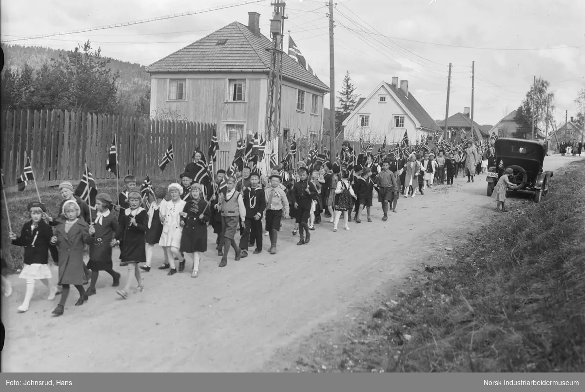 17. mai feiring 1933. Barnetog i boligomårde. Barna vifter med flagg. Bil parkert lang gaten.
