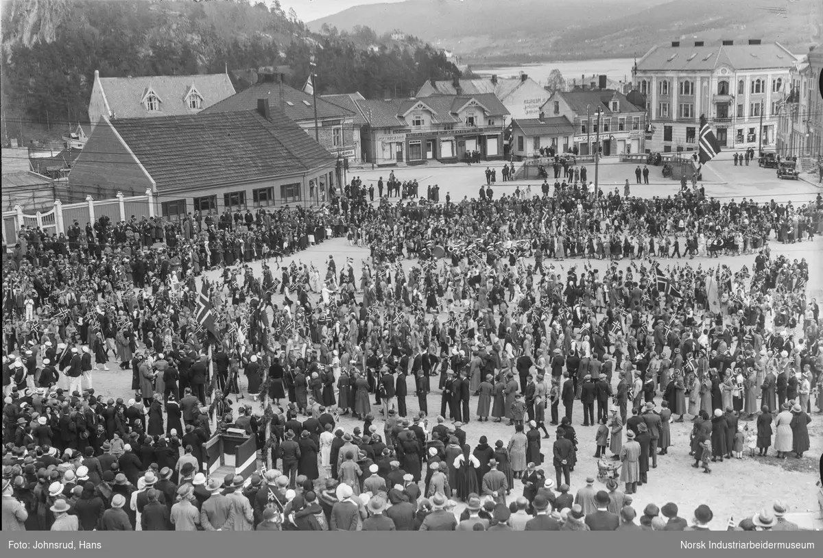 17. mai 1933. Folkemengde med flagg samlet på Notodden torg. Butikklokaler i bakgrunnen.