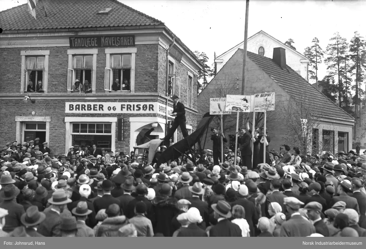 17. mai feiring 1933. Russ med lue og plakater holder tale til folkemengde på Notodden torg fra lasteplan. Folk i vinduene i hus i bakgrunnen. Butikklokale for barberer, frisør og tannlege Navelsaker.