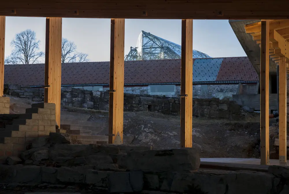 Gjennom glasset i ett av Fehns vernebygg over en middelalderruin, skimtes midtdelen av Storhamarlåven og toppen på vernebygget over domkirkeruinen.